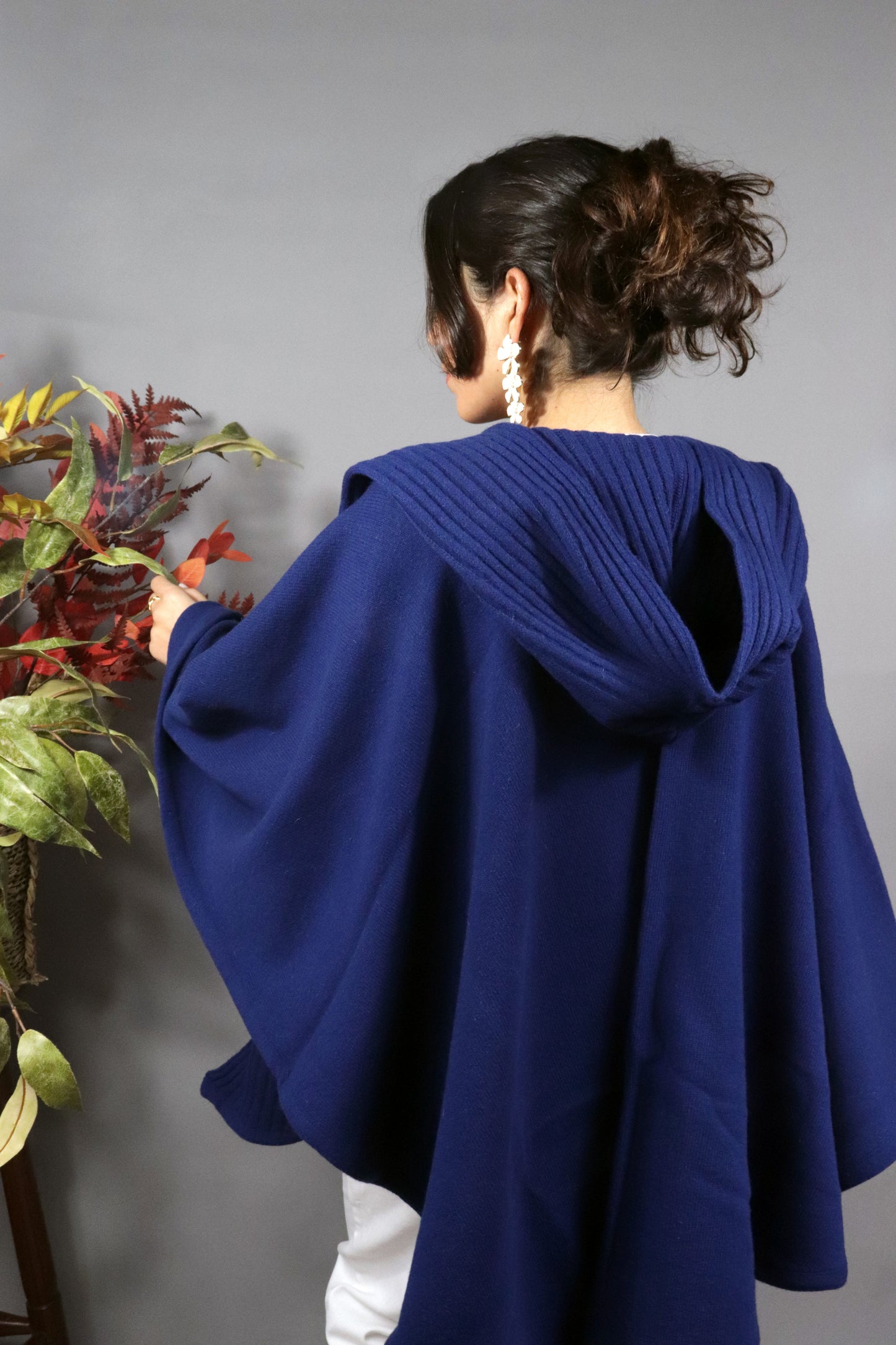 Woman seen from behind wearing a blue hooded fall cape in a neutral setting with autumn leaves decoration.