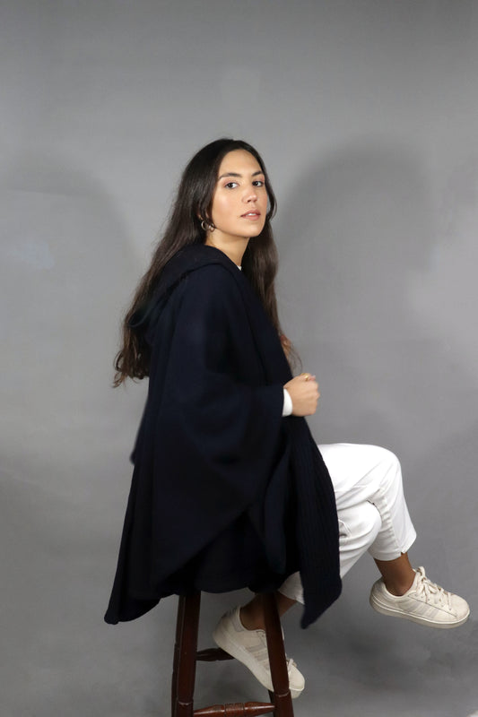 Woman seated, wearing a dark-blue hooded fall cape, paired with white pants and sneakers, against a neutral background.