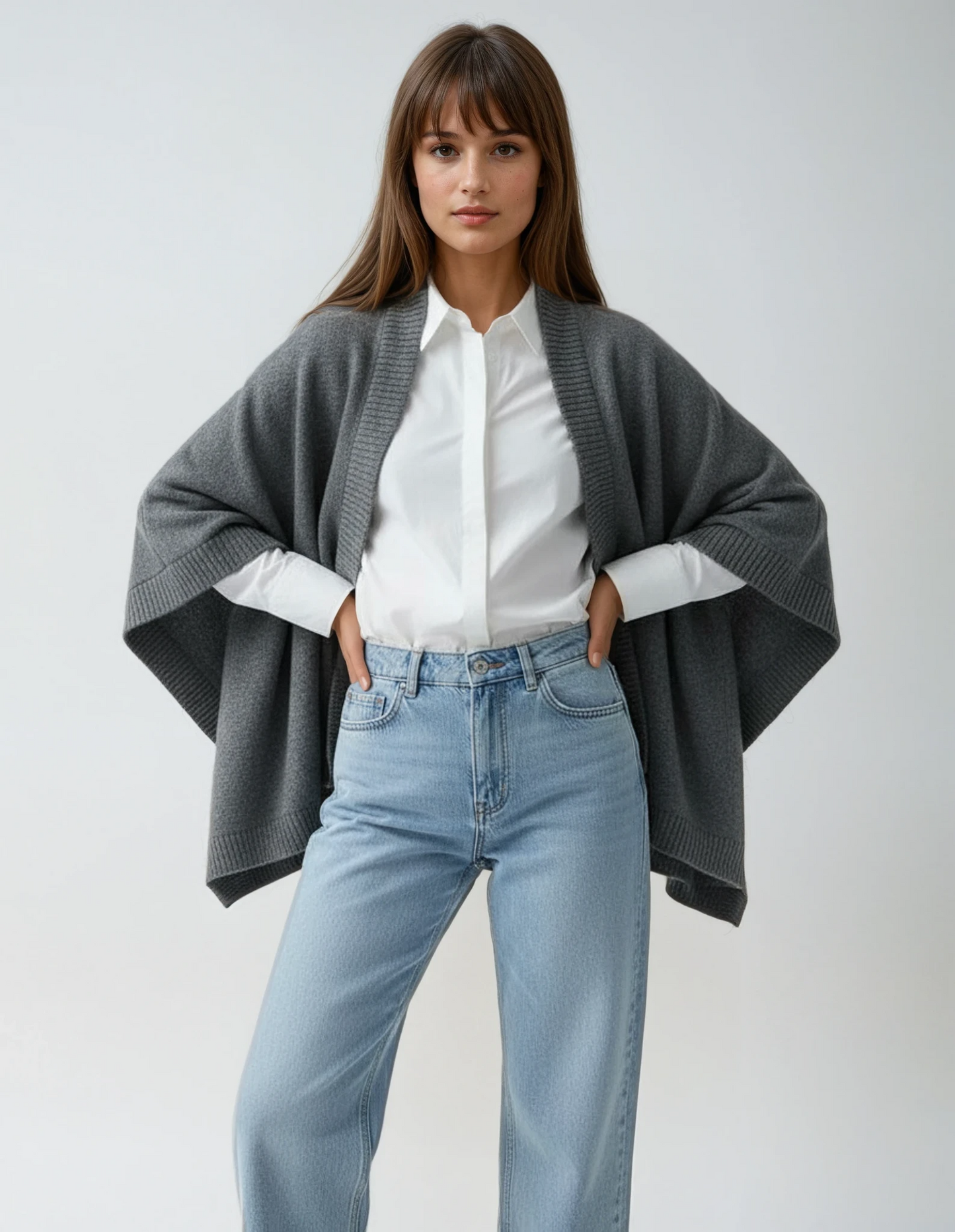 Woman wearing a dark grey cape over a white shirt and denim jeans, standing against a neutral background.
