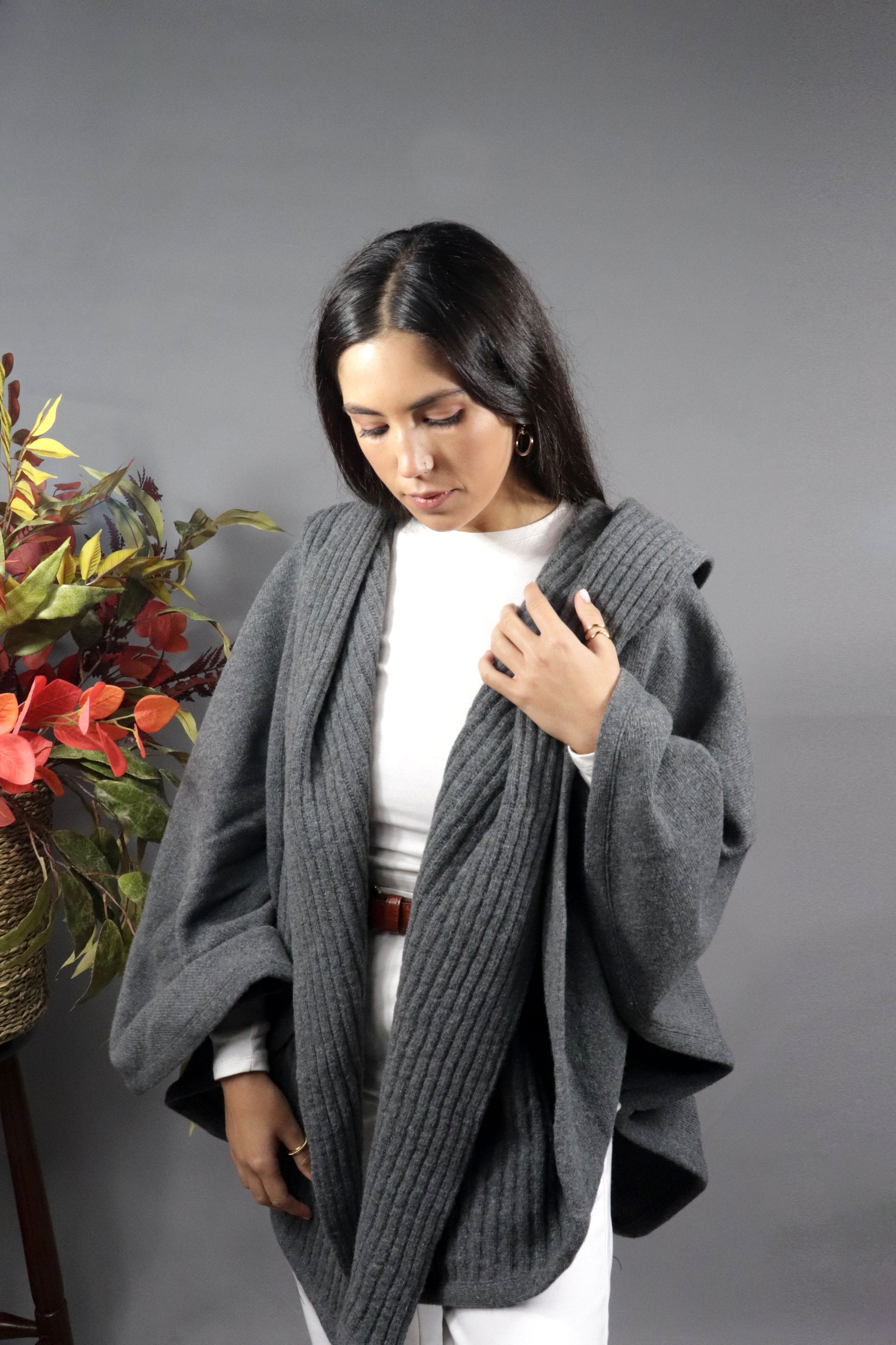 Woman seen from behind wearing a grey hooded fall cape in a neutral setting with autumn leaves decoration.