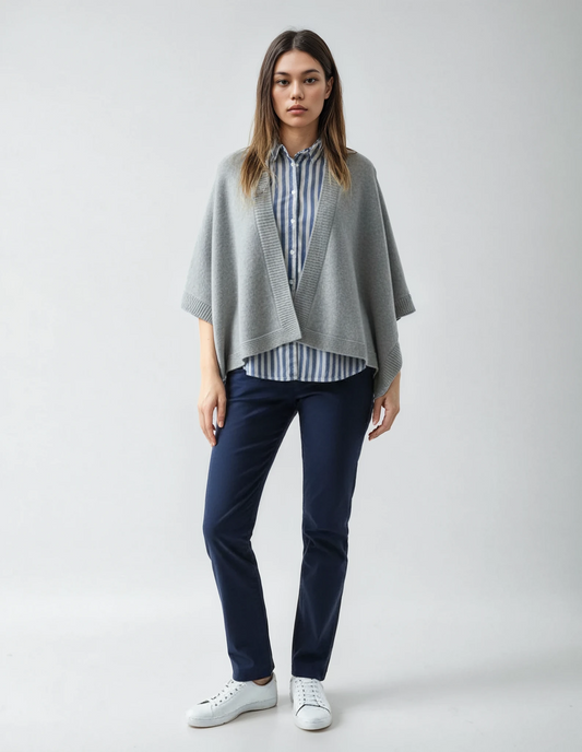Woman wearing a light grey-colored cape over a blue-and-white striped shirt and blue pants, standing against a neutral background.