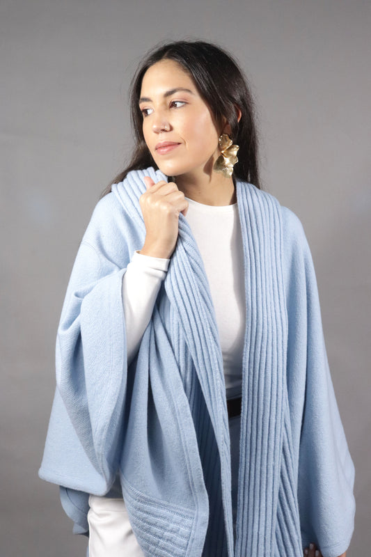 Woman standing in a light-blue hooded fall cape, styled with white clothing and gold earrings, against a neutral backdrop.