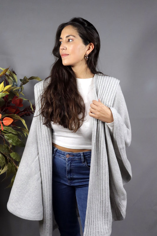 Woman seen from behind wearing a light grey hooded fall cape in a neutral setting with autumn leaves decoration.