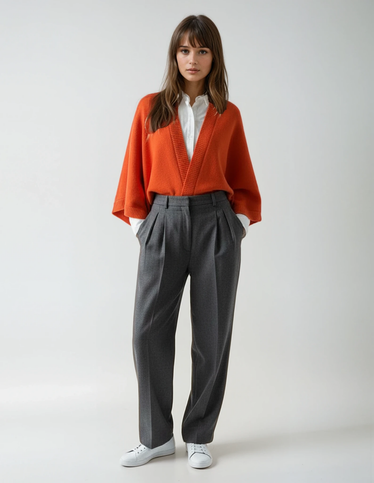 Woman wearing an orange cape over a white shirt and dark gray pants, standing against a neutral background.