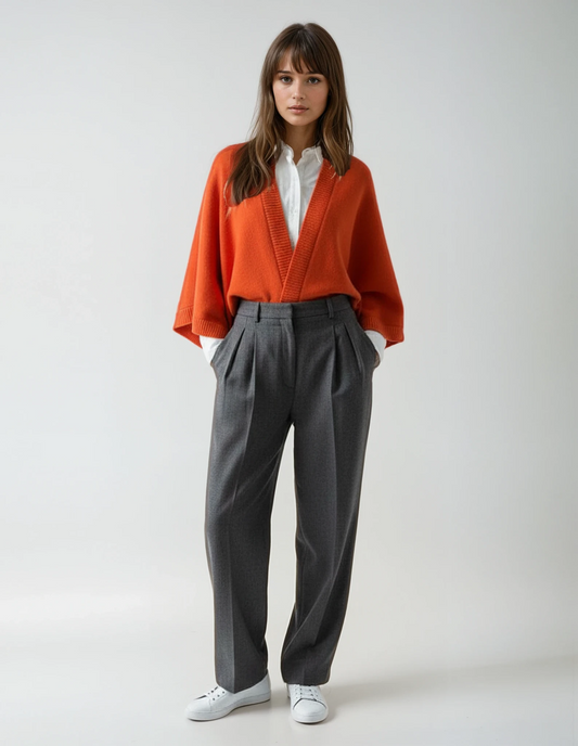 Woman wearing an orange cape over a white shirt and dark gray pants, standing against a neutral background.