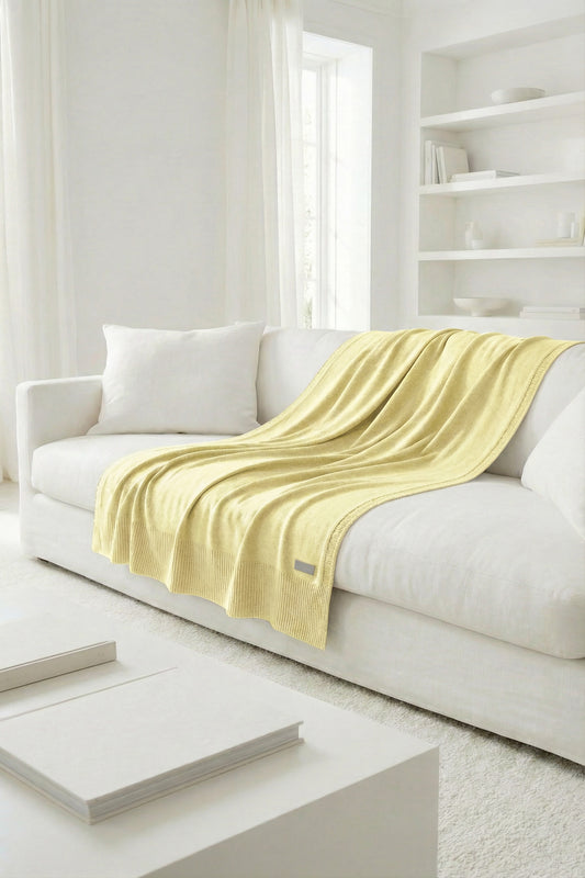 Soft yellow cotton knit throw blanket, 100% natural fiber, draped over a minimalist white couch.