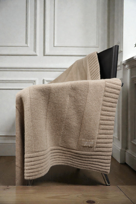 Close-up of a beige ribbed knit blanket with a textured border and brand label, draped over a dark chair.