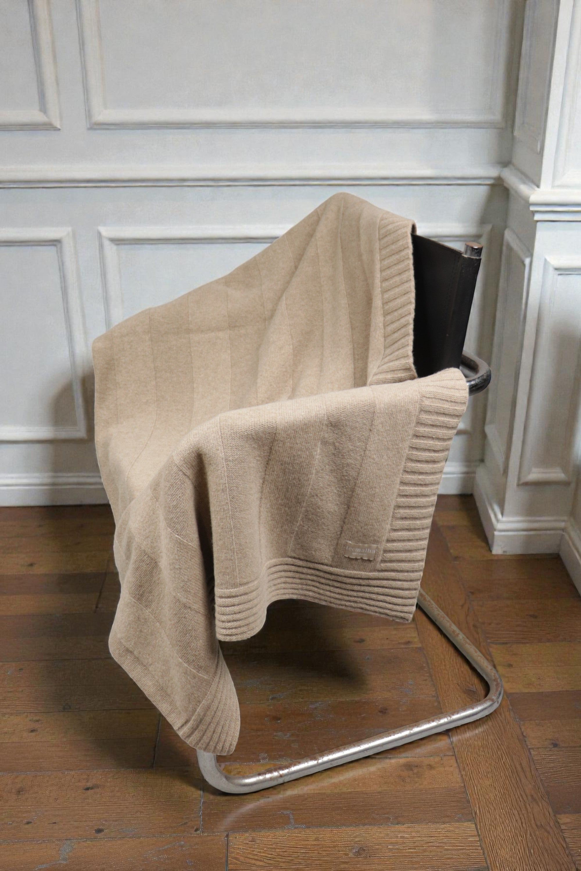 Beige ribbed knit throw blanket featuring a luxury texture and brand tag in a bright room.