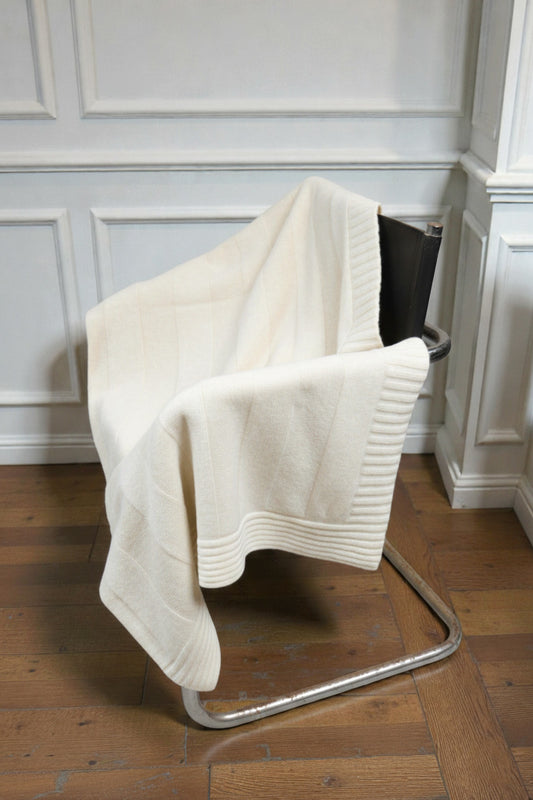 A pearl white or cream ribbed knit blanket draped over a black leather and chrome cantilever chair, set against a white paneled wall and wooden flooring.