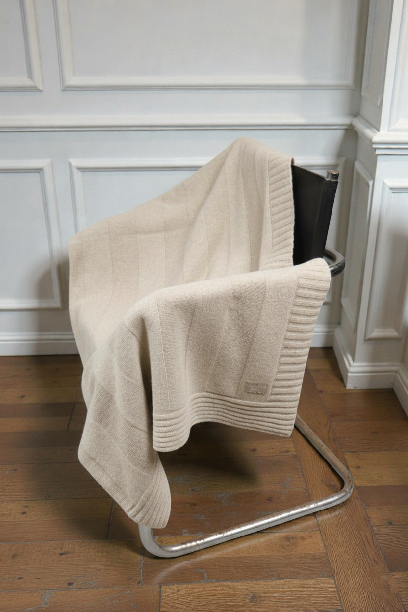 A sand ribbed knit blanket draped over a black leather and chrome cantilever chair, set against a white paneled wall and wooden flooring.