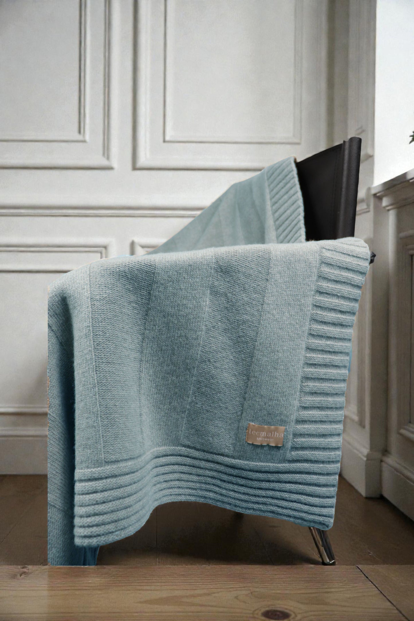 Close-up of a sapphire blue ribbed knit blanket draped over a modern black chair, showcasing the thick textured border and a small tan brand label.