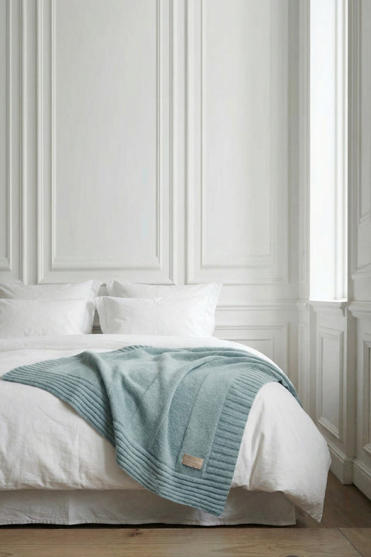 A sapphire blue ribbed knit throw blanket draped over a white bed with plush pillows against a white paneled wall.