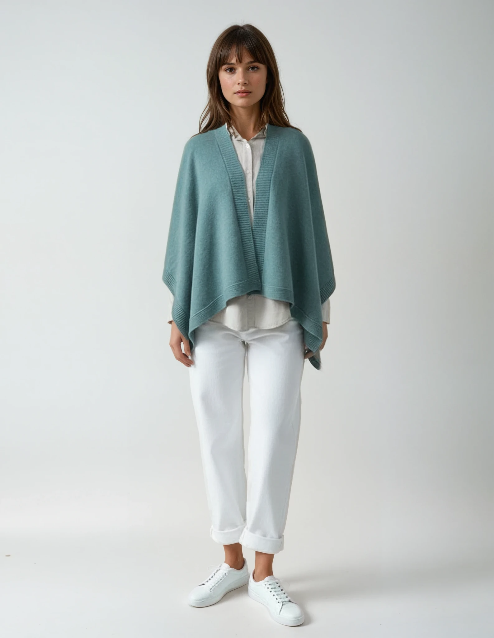 Woman wearing a sapphire blue cape over a white shirt and white pants, standing against a neutral background.