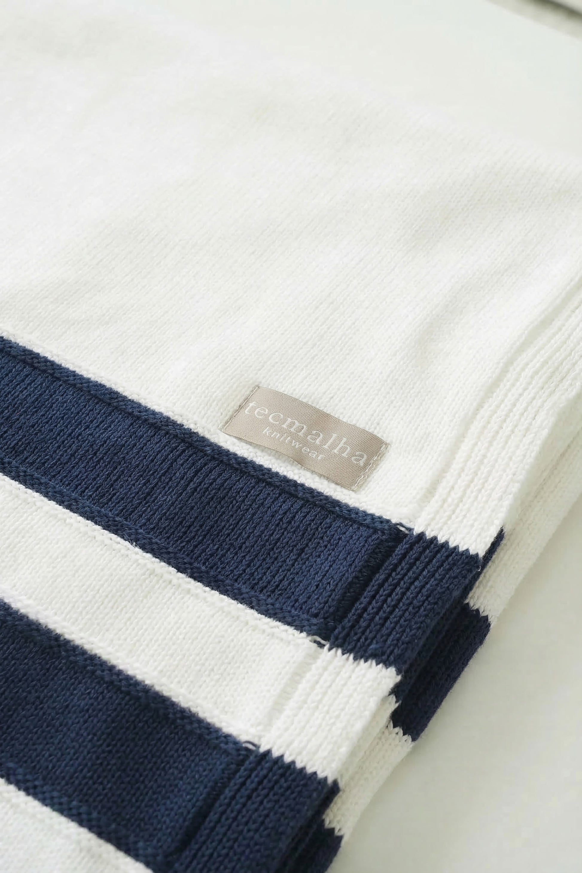 Close-up of a white knit cotton blanket showing the navy blue stripe texture and the Tecmalha brand label.