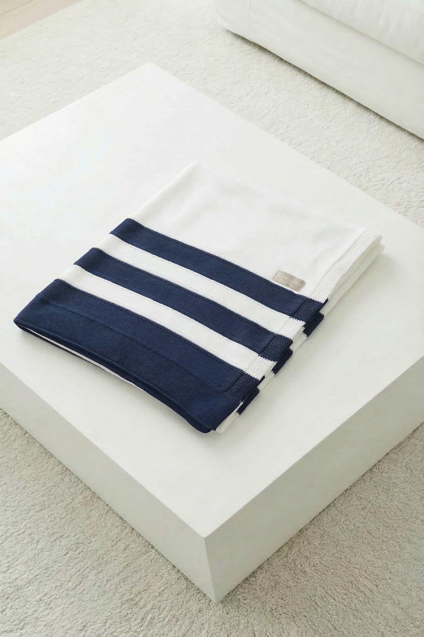 Folded white knit blanket with navy blue stripes on a minimalist white surface, showing the clean design.